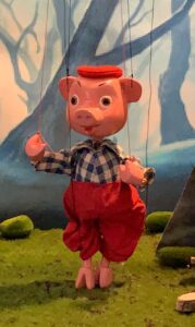 Pig marionette dressed in black and white plaid shirt and red pants with suspenders